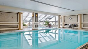 Une piscine intérieure avec des parois en verre transparent, un sol carrelé et un espace détente avec des tables et des chaises.