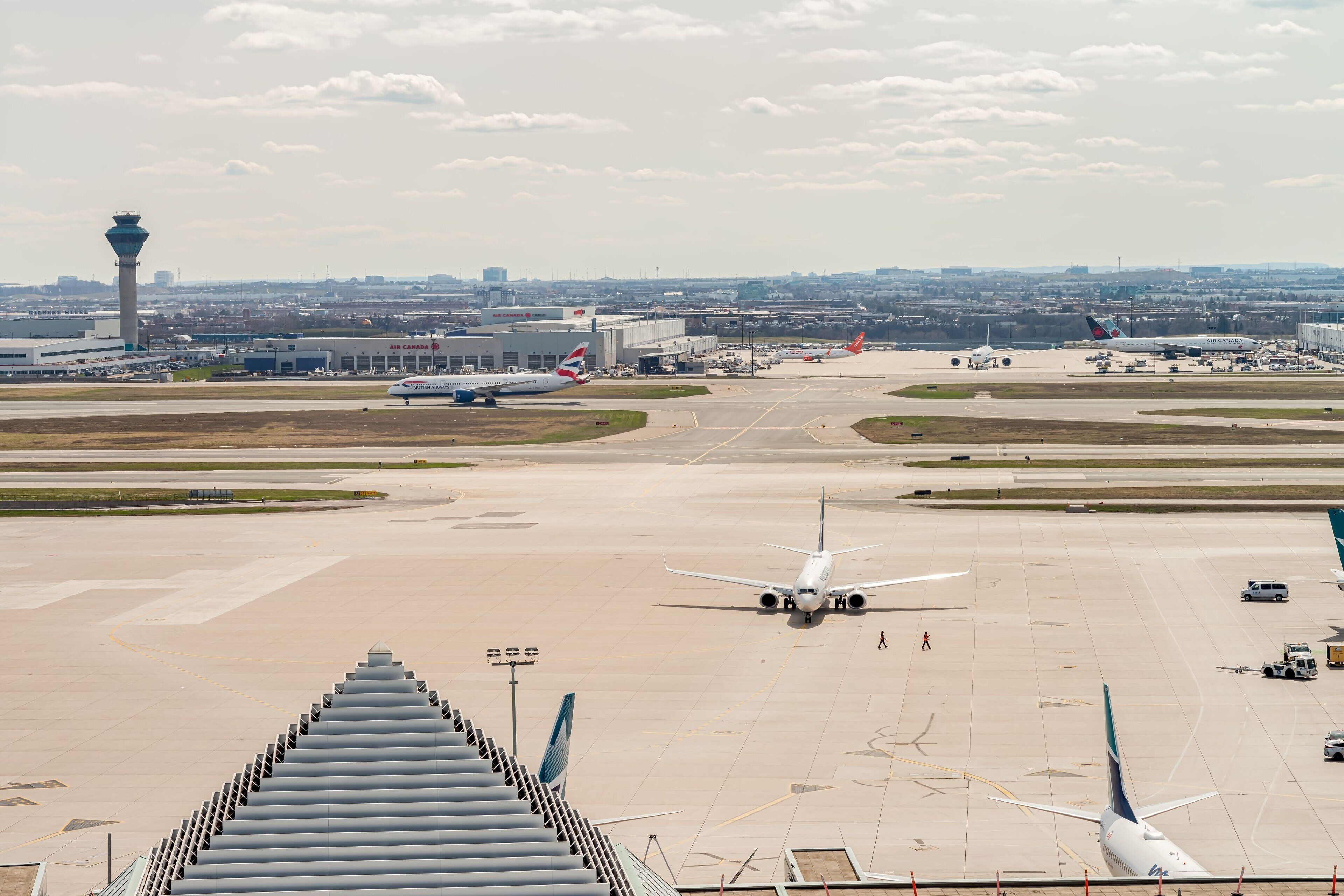 Una pista de aeropuerto con múltiples aviones, una torre de control y edificios terminales al fondo.