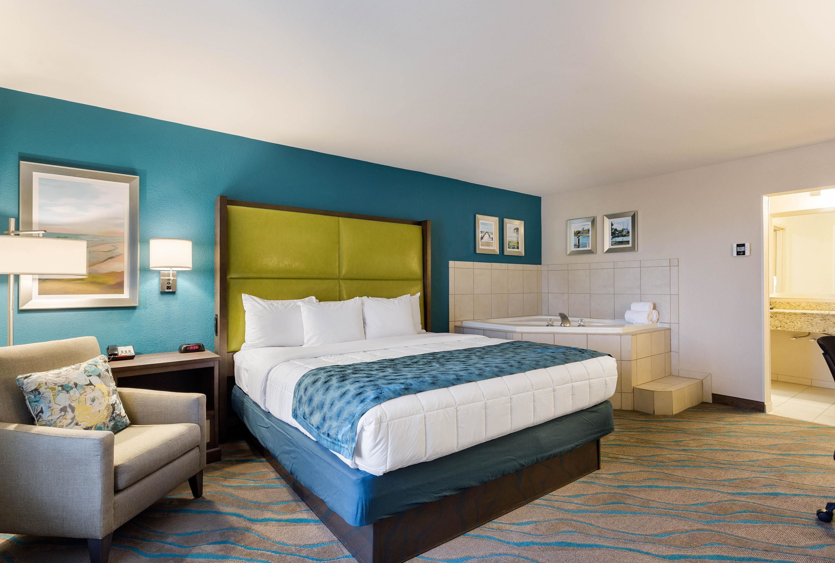 A hotel room with a large bed, a chair, a nightstand, a lamp, a painting, a bathroom with a bathtub, and a sink.