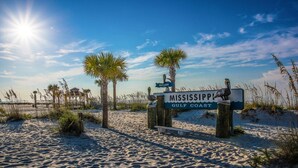 Un letrero de playa con la inscripción 'Mississippi Gulf Coast', rodeado de arena y palmeras.