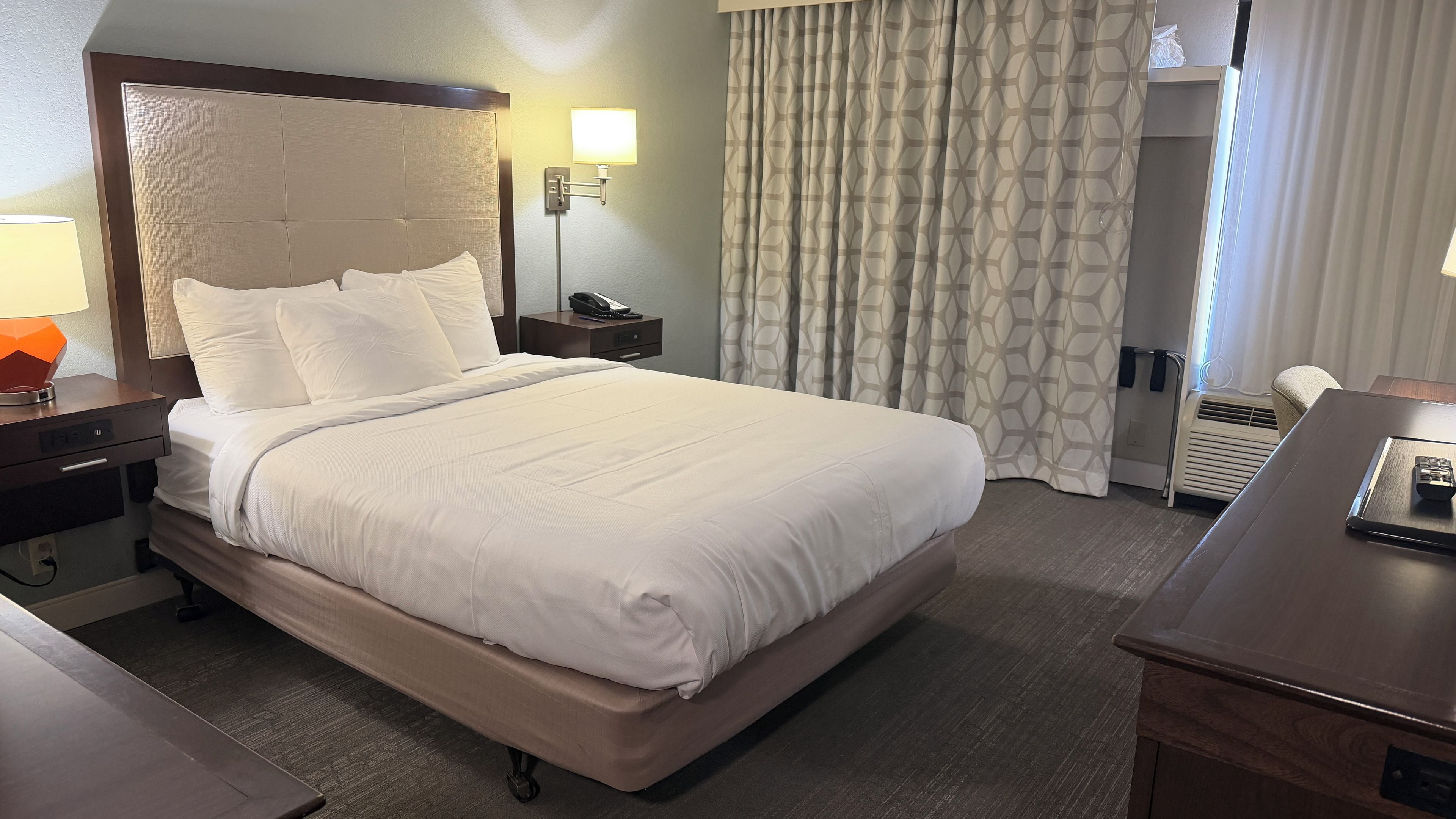 Quarto de hotel com uma cama grande, mesa de cabeceira com abajur, escrivaninha com controle remoto e janela com cortinas.