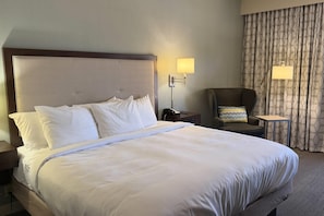 A hotel room with a large bed, a nightstand, a chair, a desk, and a lamp.