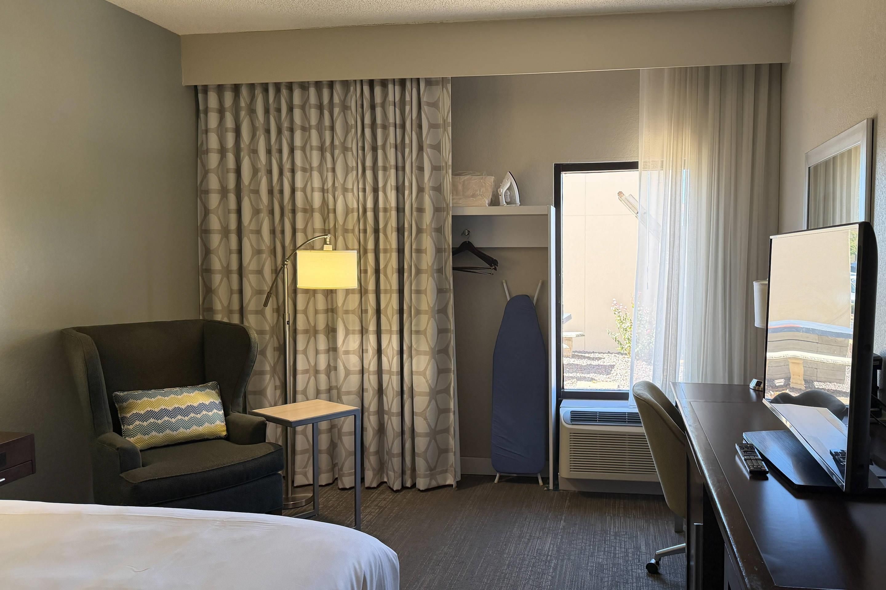 A hotel room with a bed, a desk with a computer, a chair, a lamp, a wardrobe, and a window with curtains.
