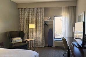 Una habitación de hotel con una cama, un escritorio con computadora, una silla, una lámpara, un armario y una ventana con cortinas.