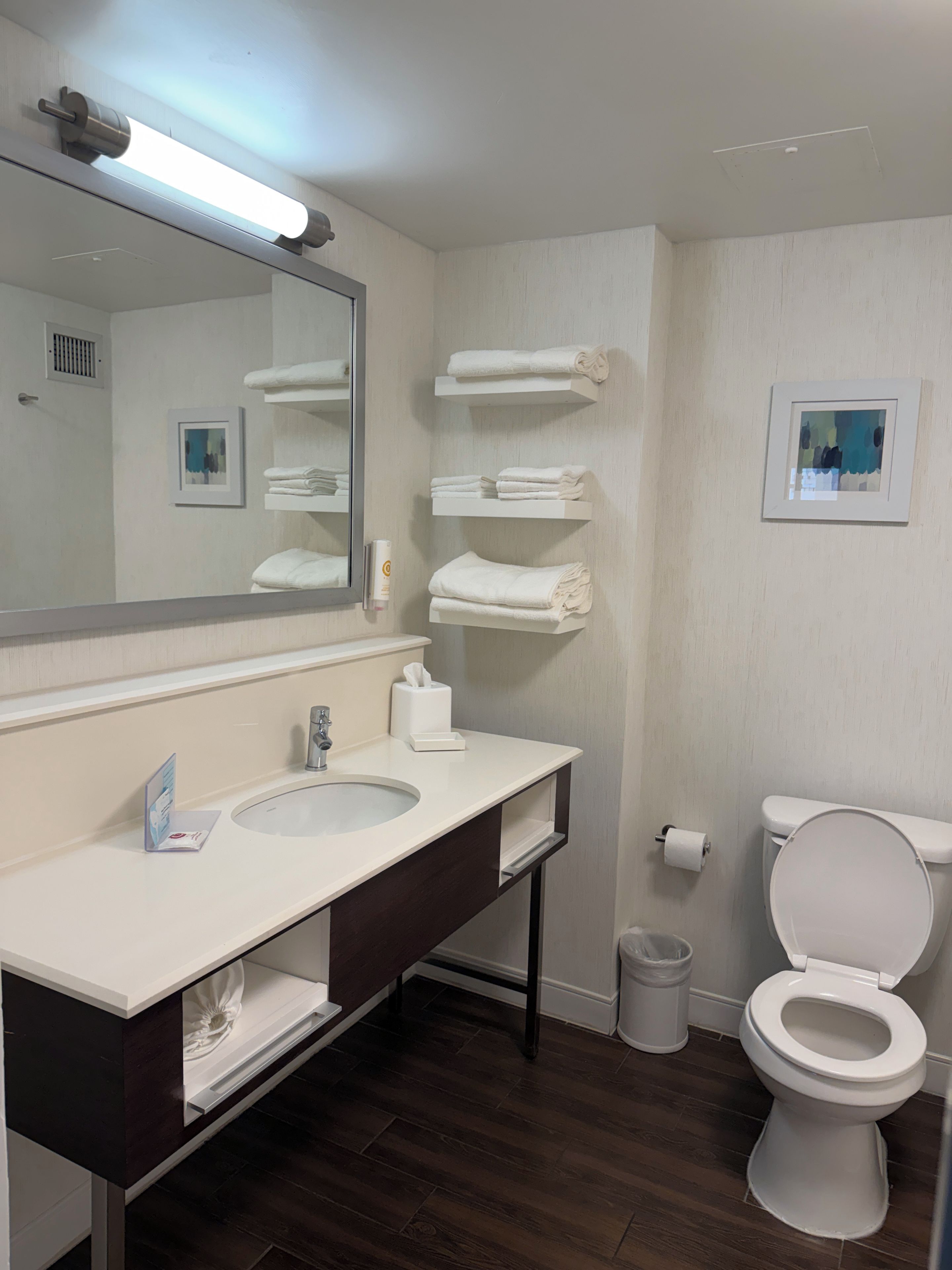 A bathroom with a white sink, mirror, and toilet. There are towels on a shelf and a trash can.