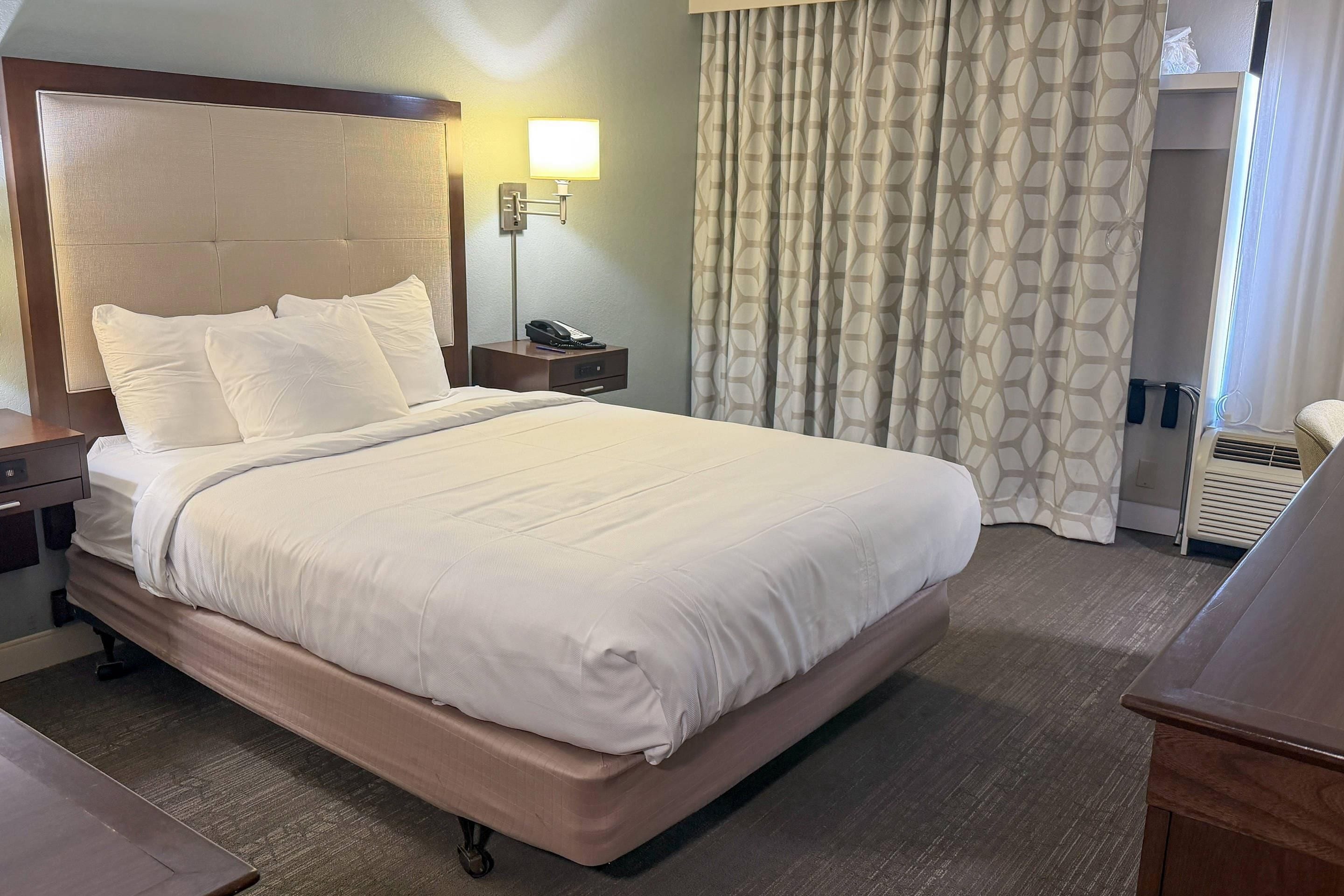 Habitación de hotel con una cama grande, una mesita de noche con lámpara, teléfono y ventana con cortinas.