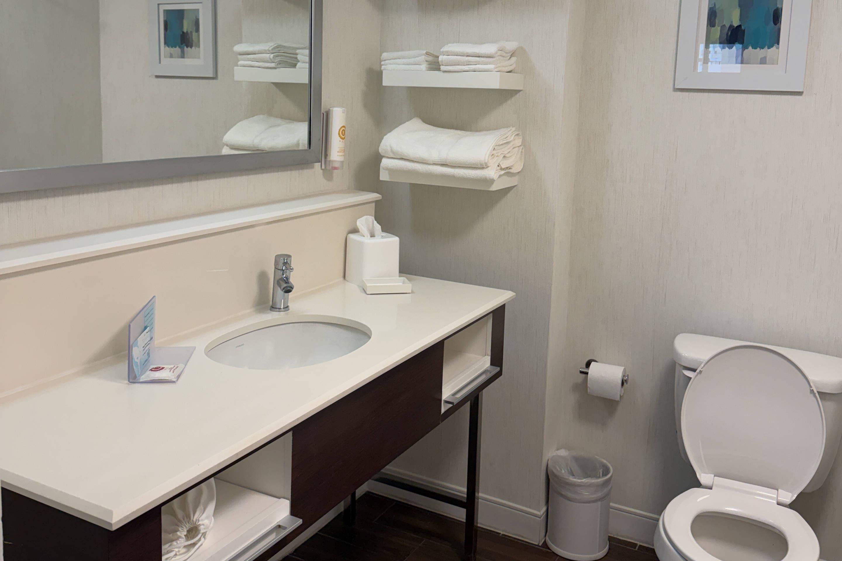 Une salle de bain avec un lavabo blanc, un miroir et des toilettes. Des serviettes sont pliées et empilées sur une étagère au-dessus des toilettes.