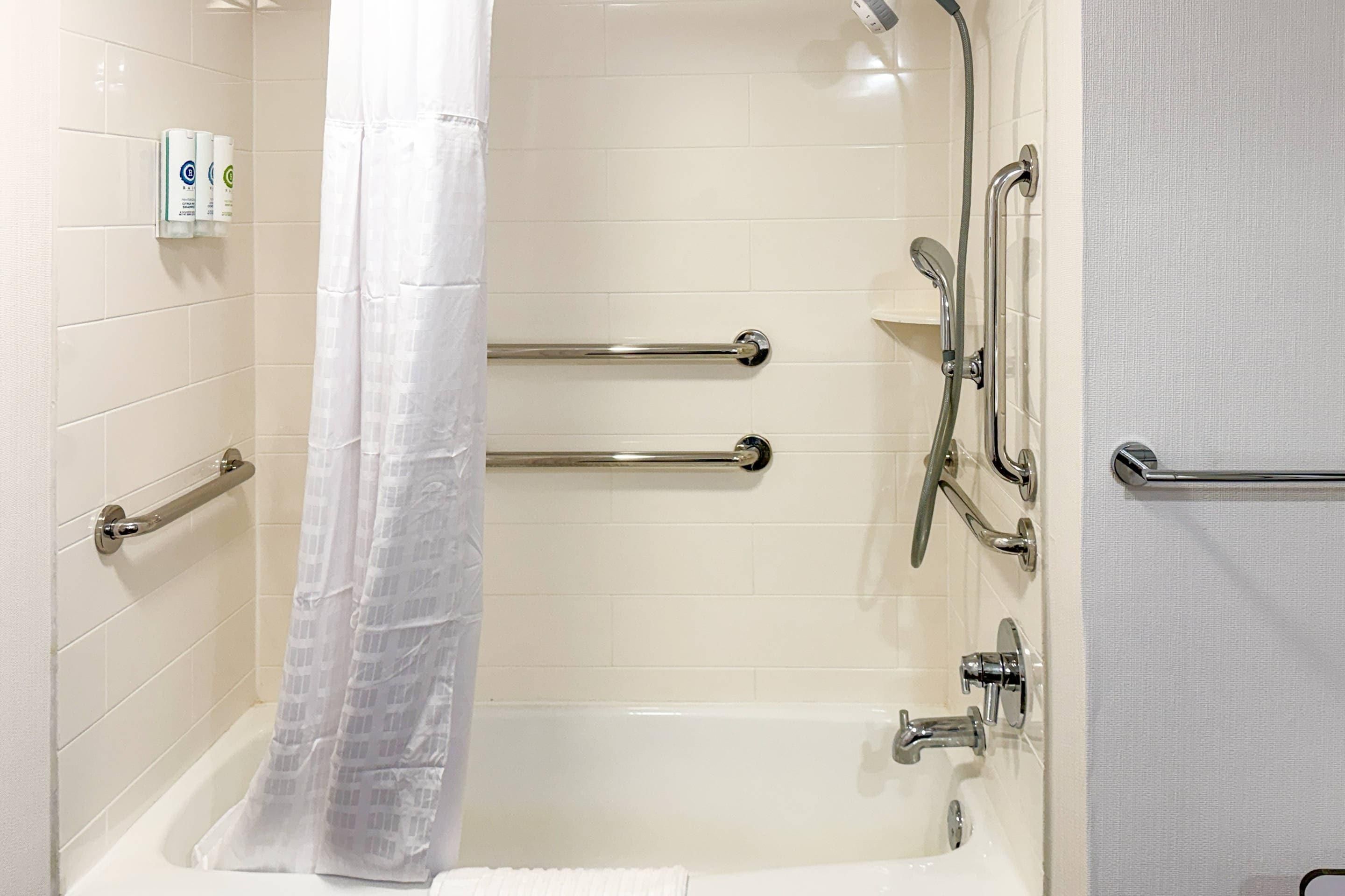 A bathroom with a bathtub, shower, and handrails.