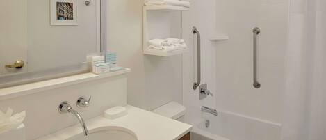 A modern bathroom with a sink, toilet, and shower. There are white towels on a shelf, a handrail, and a picture on the wall.