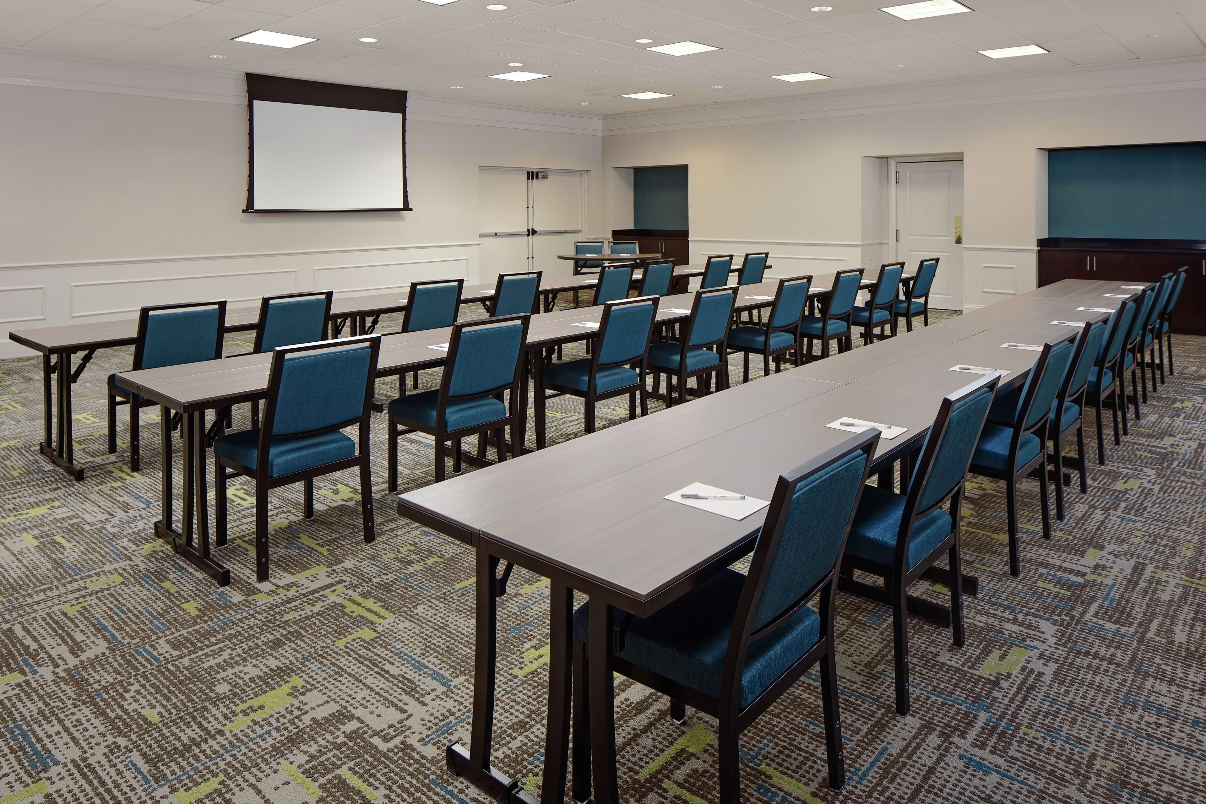 A conference room with multiple tables and chairs, a projector screen, and a carpeted floor.