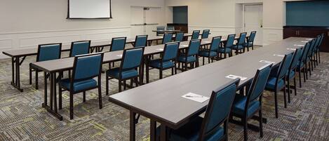 A conference room with multiple tables and chairs, a projector screen, and a carpeted floor.