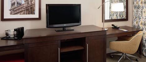 A hotel room with a dark wooden desk, a television, a red suitcase, a chair, and a framed picture of a building.