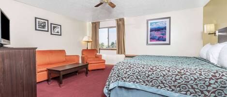 A hotel room with a bed, a sofa, a chair, a desk, a ceiling fan, and a window with curtains.