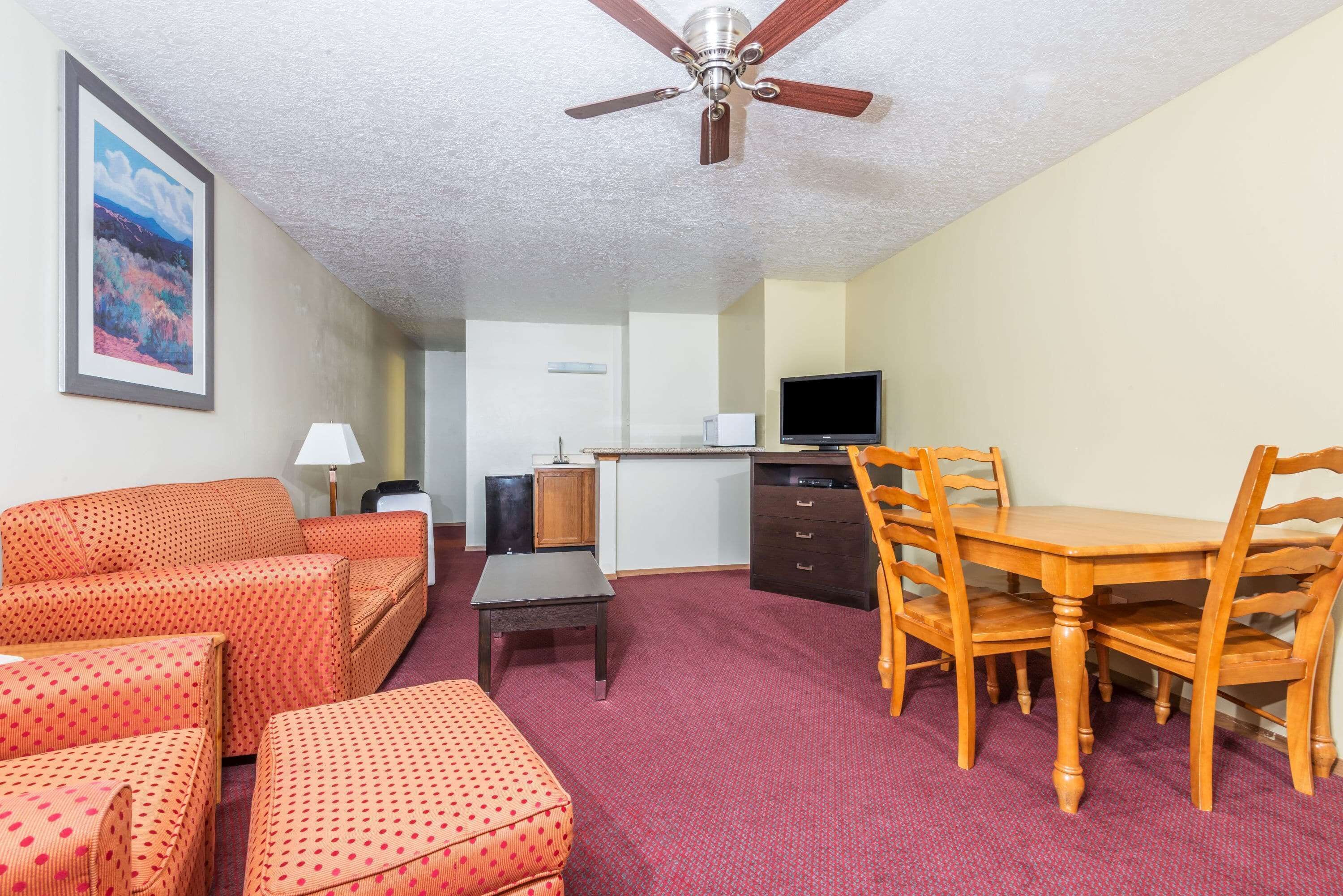 A compact hotel room with a sofa, ottoman, wooden dining table, chairs, a TV, and a ceiling fan.