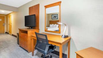 Habitación de hotel con un escritorio de madera, una silla de oficina negra, un televisor sobre un mueble de madera, una lámpara de noche y un espejo.