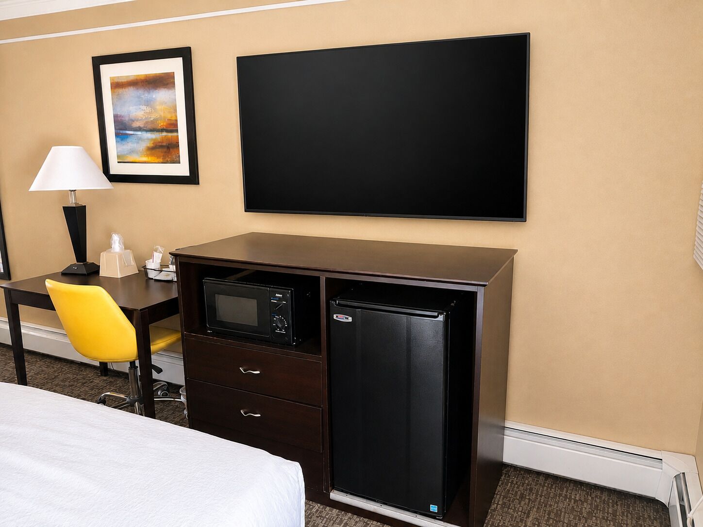 A hotel room with a bed, a desk with a lamp, a microwave, a refrigerator, a chair, and a framed picture on the wall.