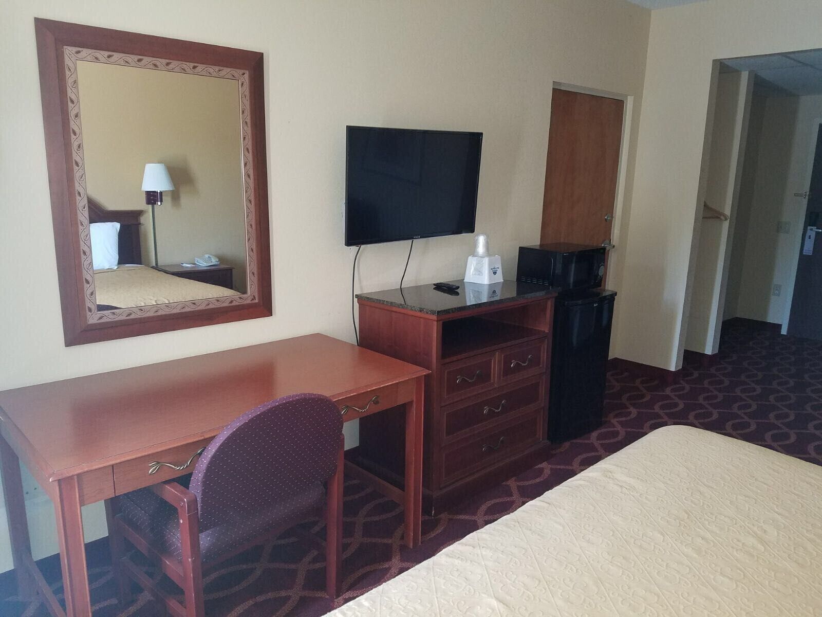 Habitación de hotel con cama, escritorio de madera, silla, cómoda, televisor, microondas y refrigerador pequeño.