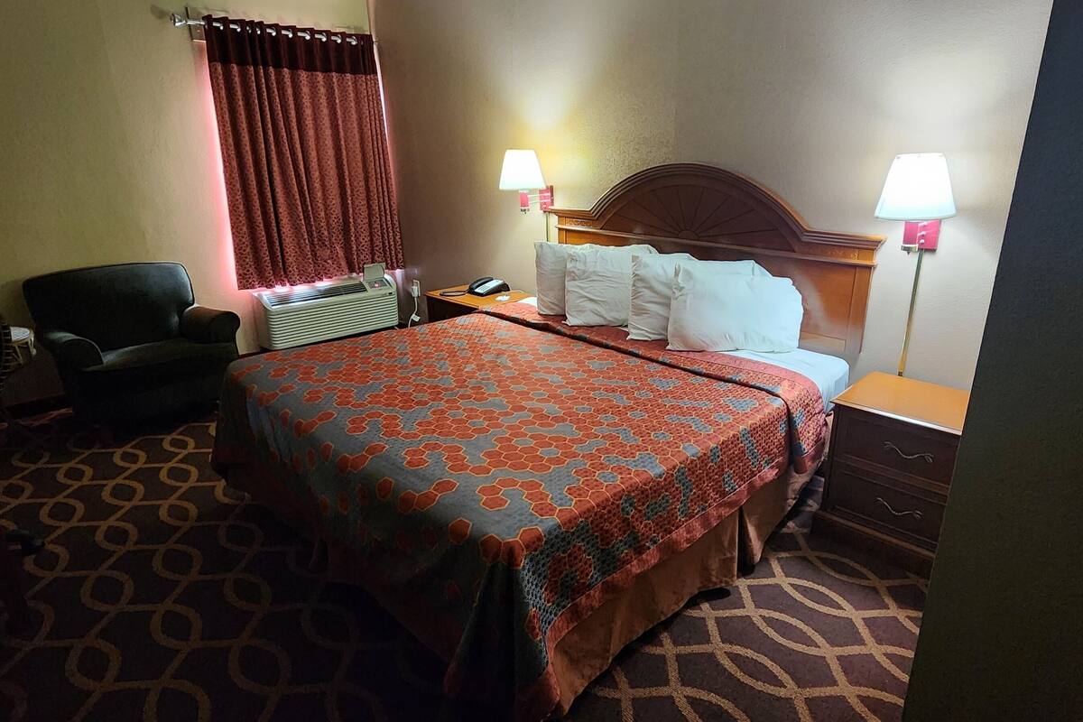 A hotel room with a bed, two bedside lamps, a chair, a desk, and a heater.