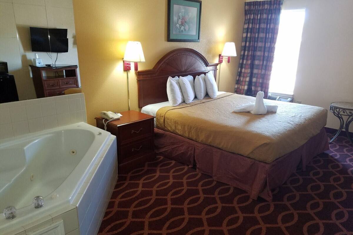 A hotel room with a large bed, two bedside lamps, a flat-screen TV, a bathroom with a bathtub, and a patterned carpet.