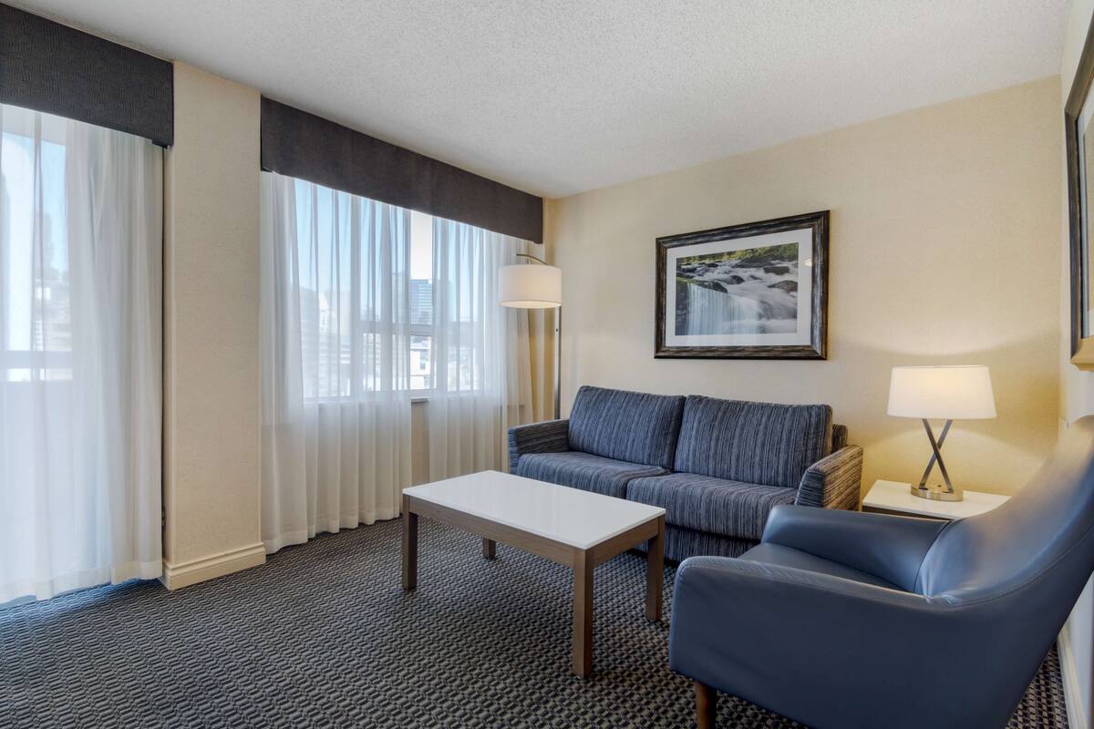 Ein Hotelzimmer mit Sofa, Sessel, Tisch, Lampe, einem gerahmten Bild an der Wand und einem Fenster mit durchscheinenden Vorhängen.