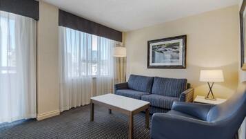 Habitación de hotel con sofá, una silla, una mesa, una lámpara, un cuadro enmarcado en la pared y una ventana con cortinas translúcidas.