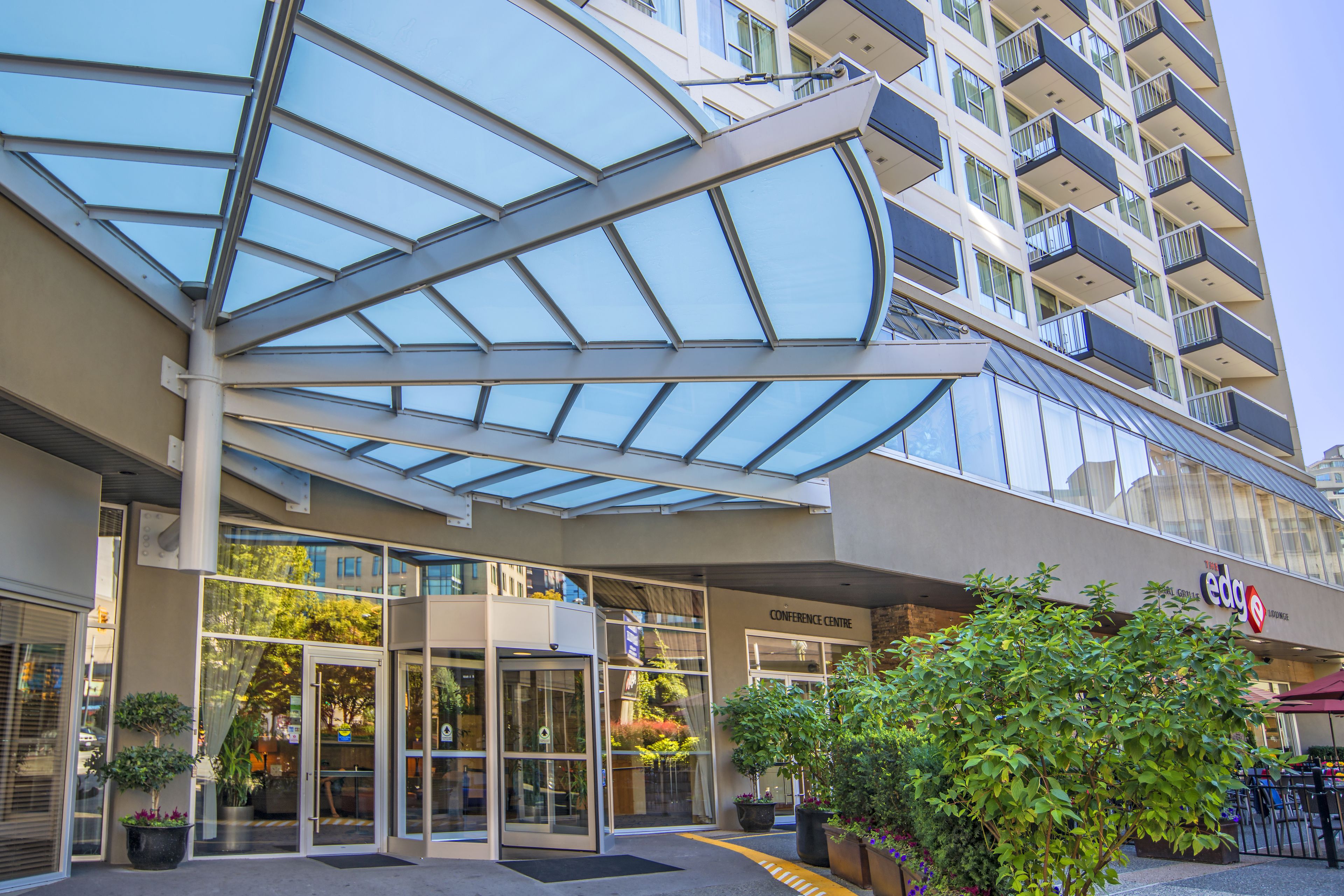 L’entrée d’un hôtel, avec un auvent en verre, une enseigne indiquant le centre de conférences et des plantes en pot.