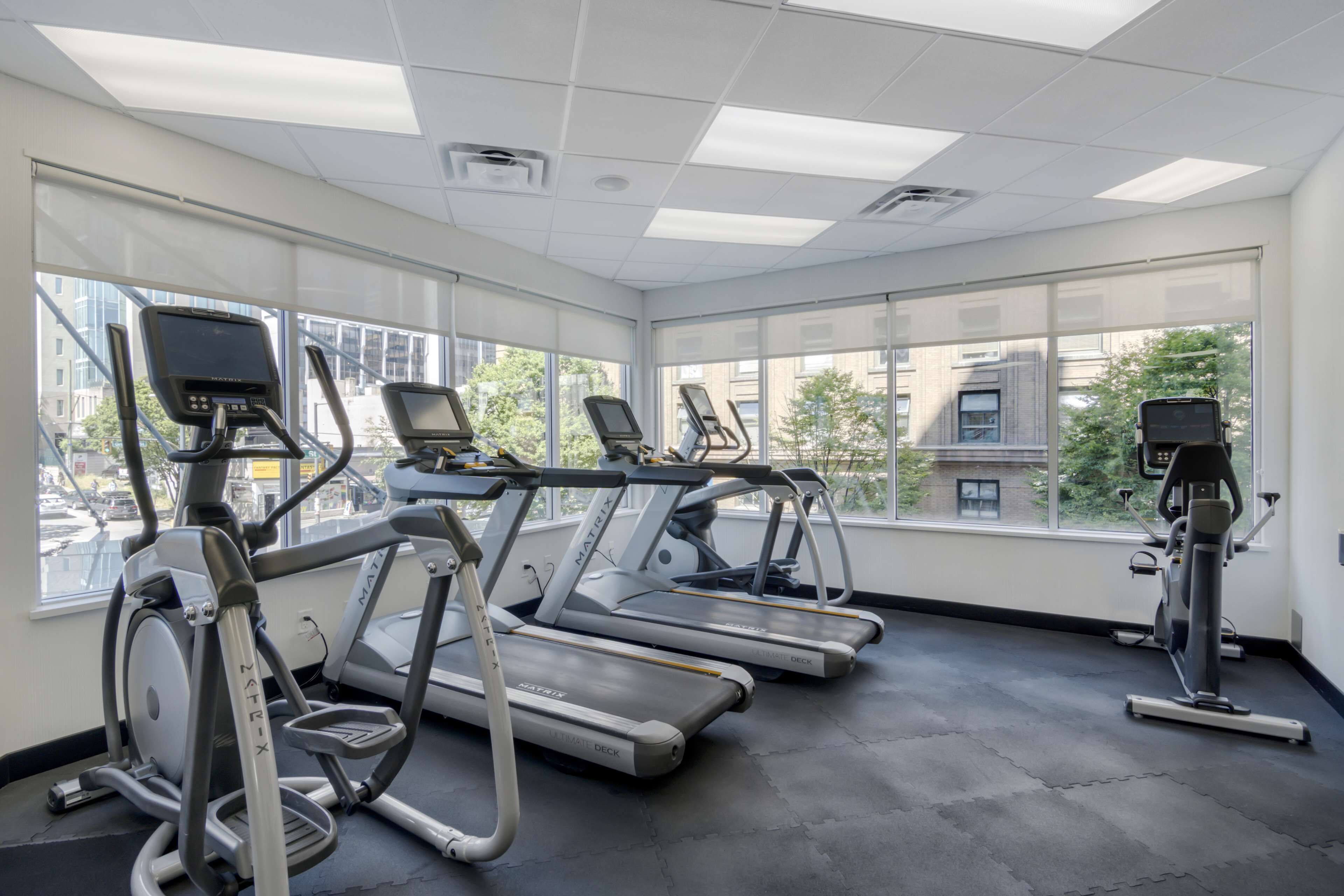 A gym with treadmills and elliptical machines, large windows, and a view of buildings and trees.