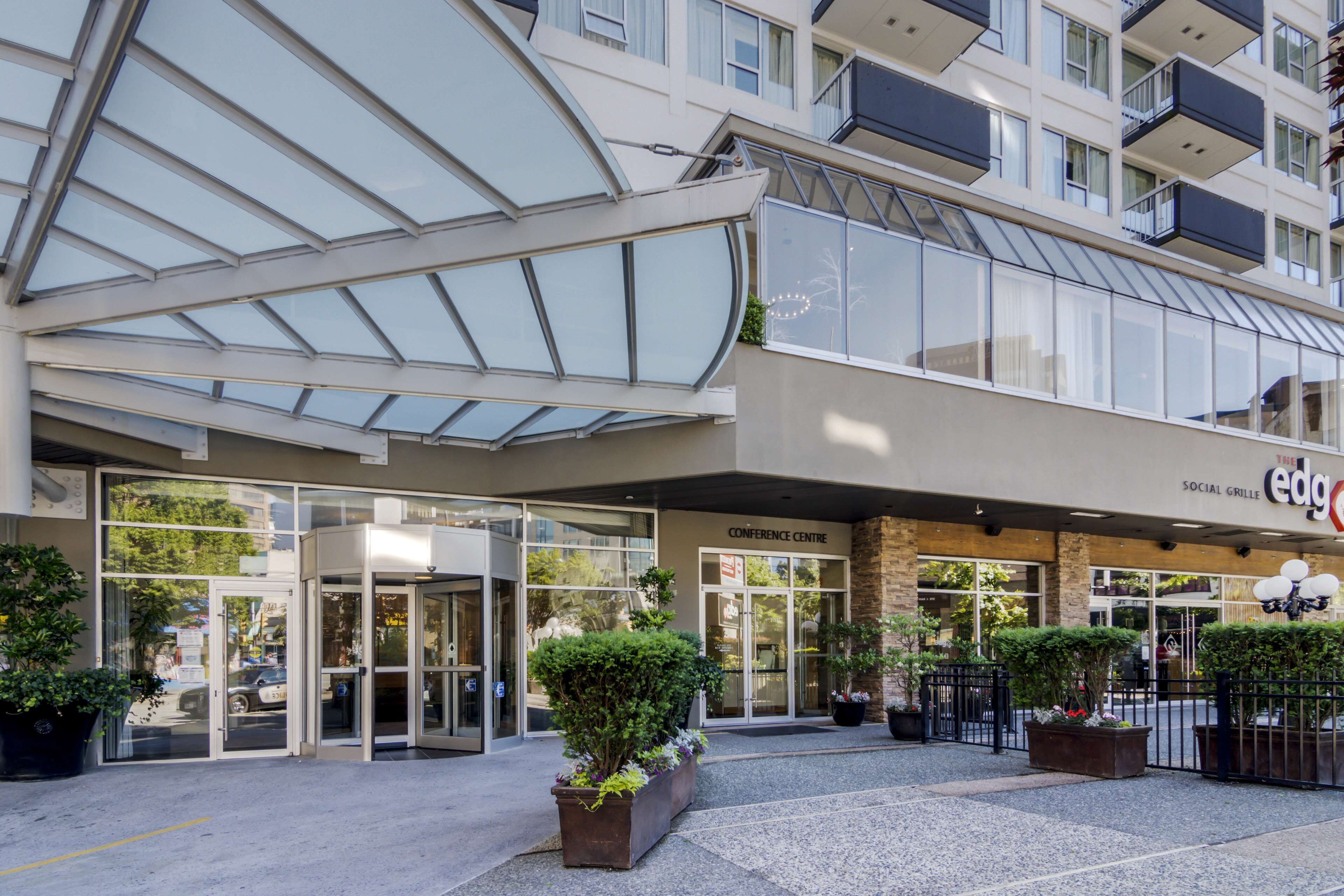 Une entrée d’hôtel moderne avec une façade en verre, une enseigne indiquant la présence d’un centre de conférences et des plantes en pot.