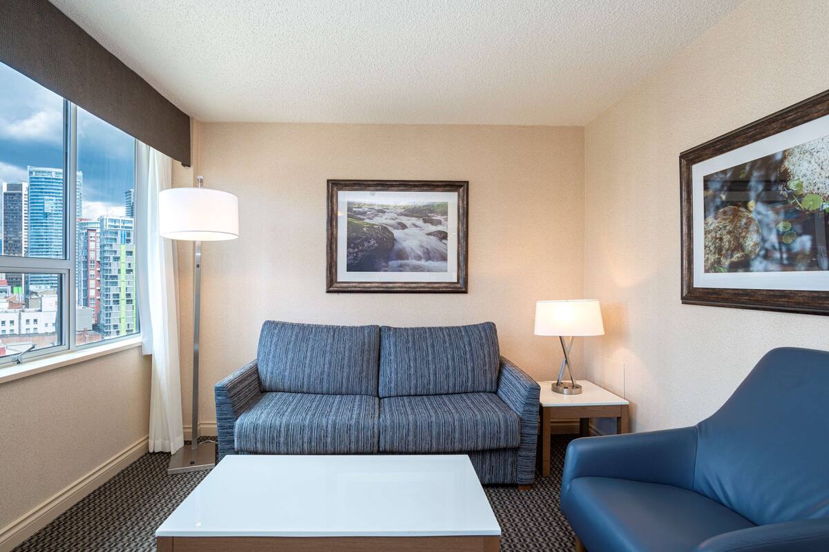 Ein Hotelzimmer mit großem Fenster, einer Couch, einem Sessel, einem Tisch und zwei Lampen.