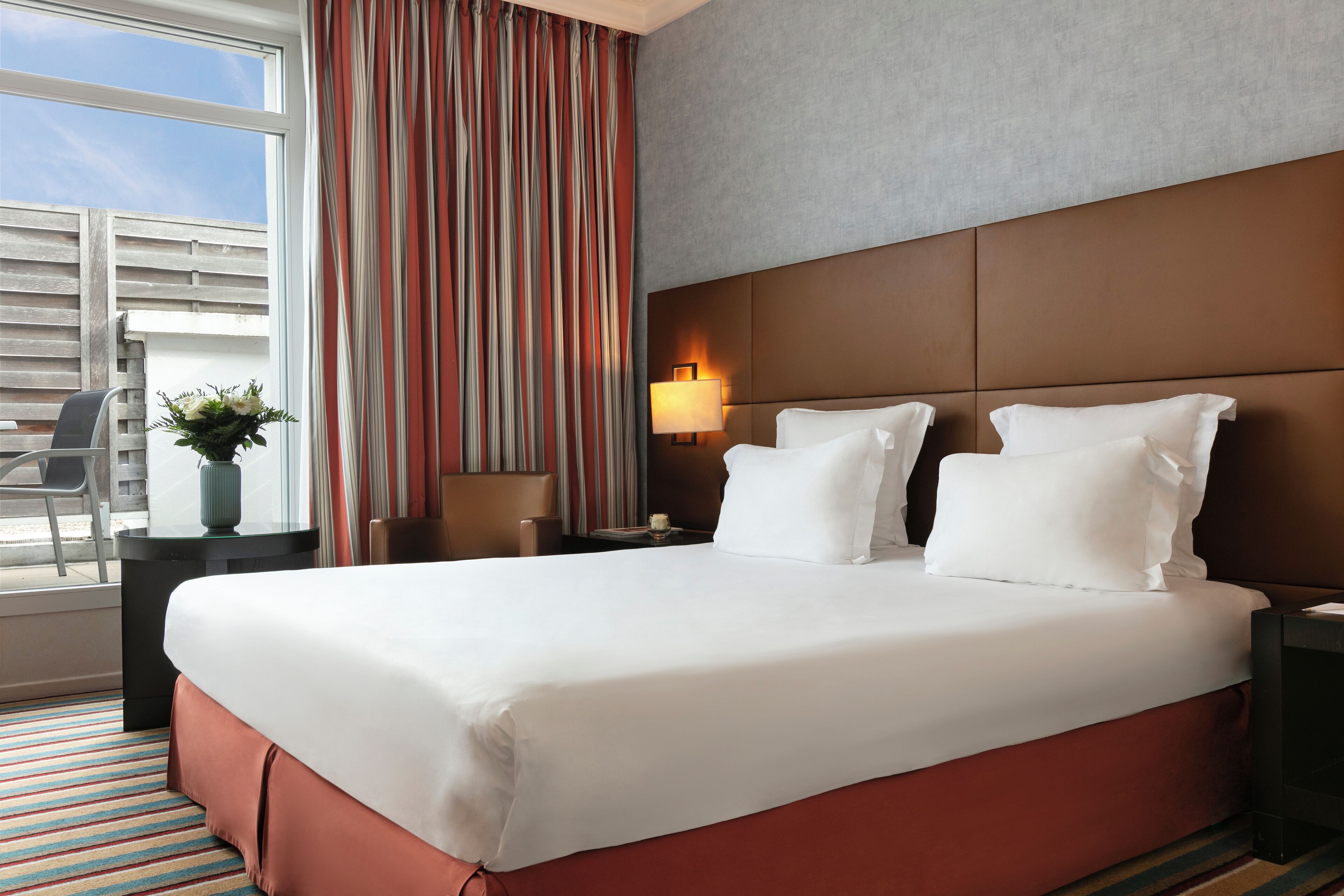 Une chambre d’hôtel avec un grand lit, une table de chevet avec une lampe, une chaise et une fenêtre avec des rideaux.