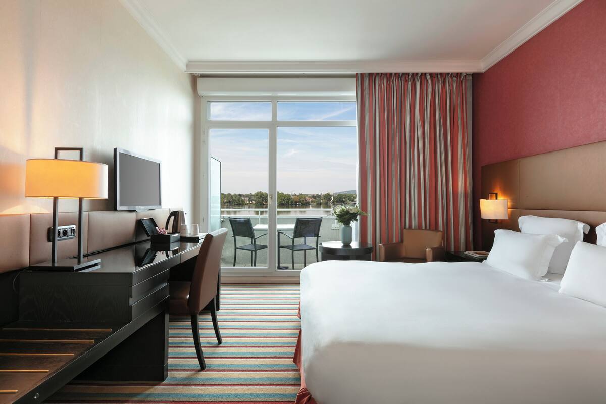 Quarto de hotel com cama, escrivaninha, cadeiras e televisão.