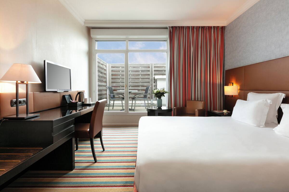 Quarto de hotel com cama, escrivaninha, cadeira, televisão e vista para o exterior.