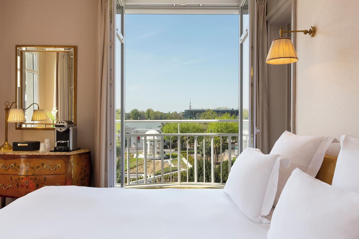 Quarto de hotel com uma cama grande, cômoda com espelho, luminária e vista para a cidade e áreas verdes.