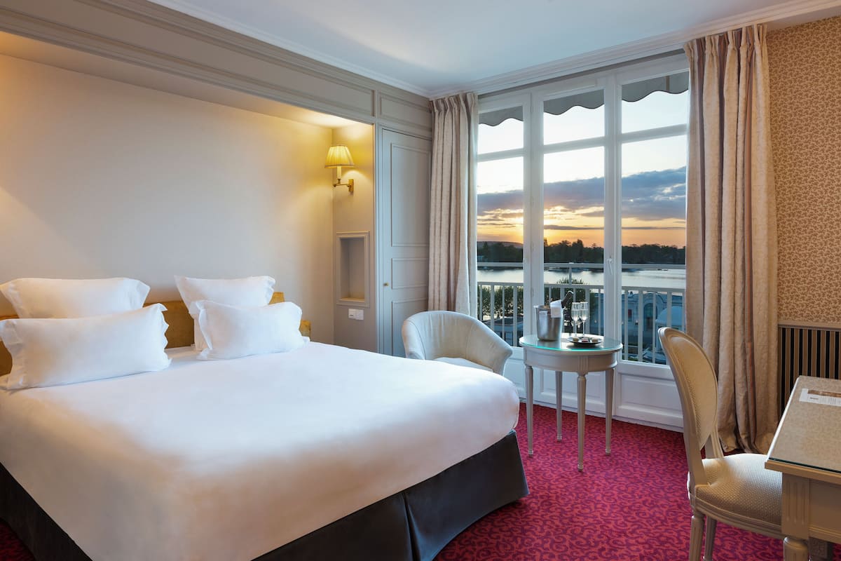 Quarto de hotel com uma cama grande, uma mesinha com um copo, uma cadeira e vista para um corpo d'água ao pôr do sol.