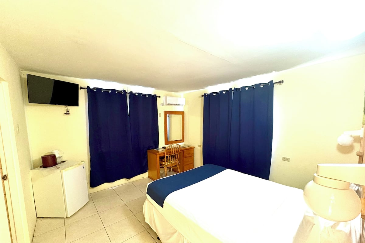 A hotel room with a bed, a desk, a chair, a dresser, a television, and a toilet.
