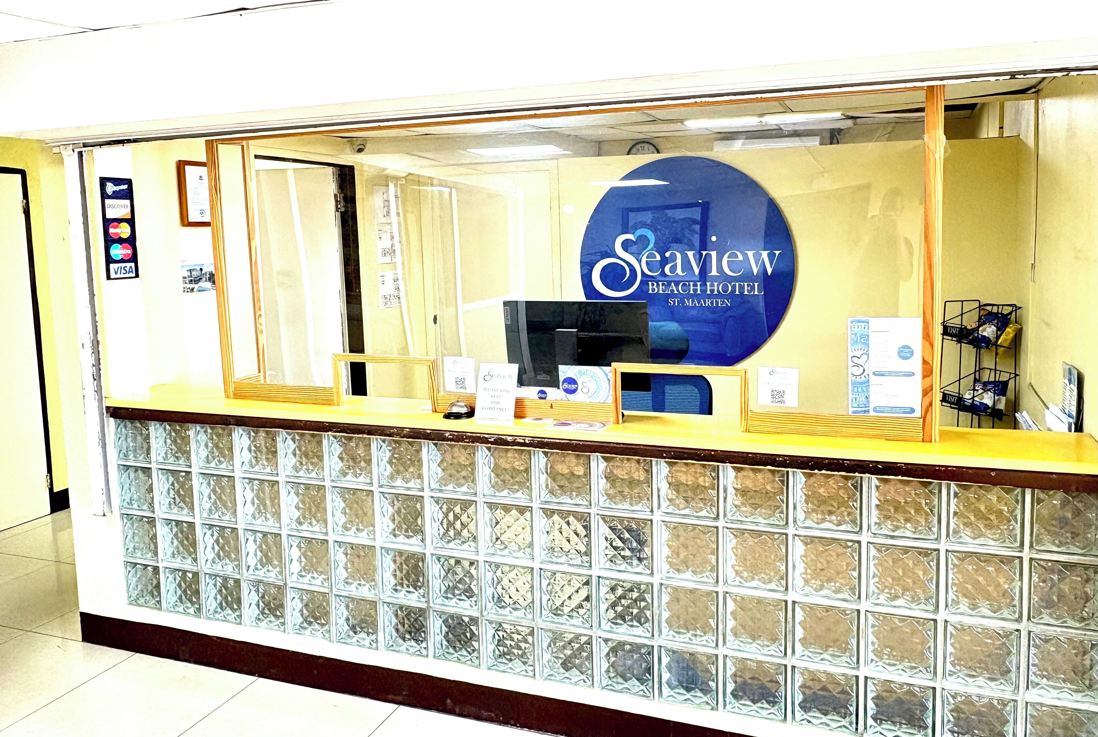 Een receptiebalie met een glazen scheidingswand, een bord met de tekst Seaview Beach Hotel en een betaalterminal.