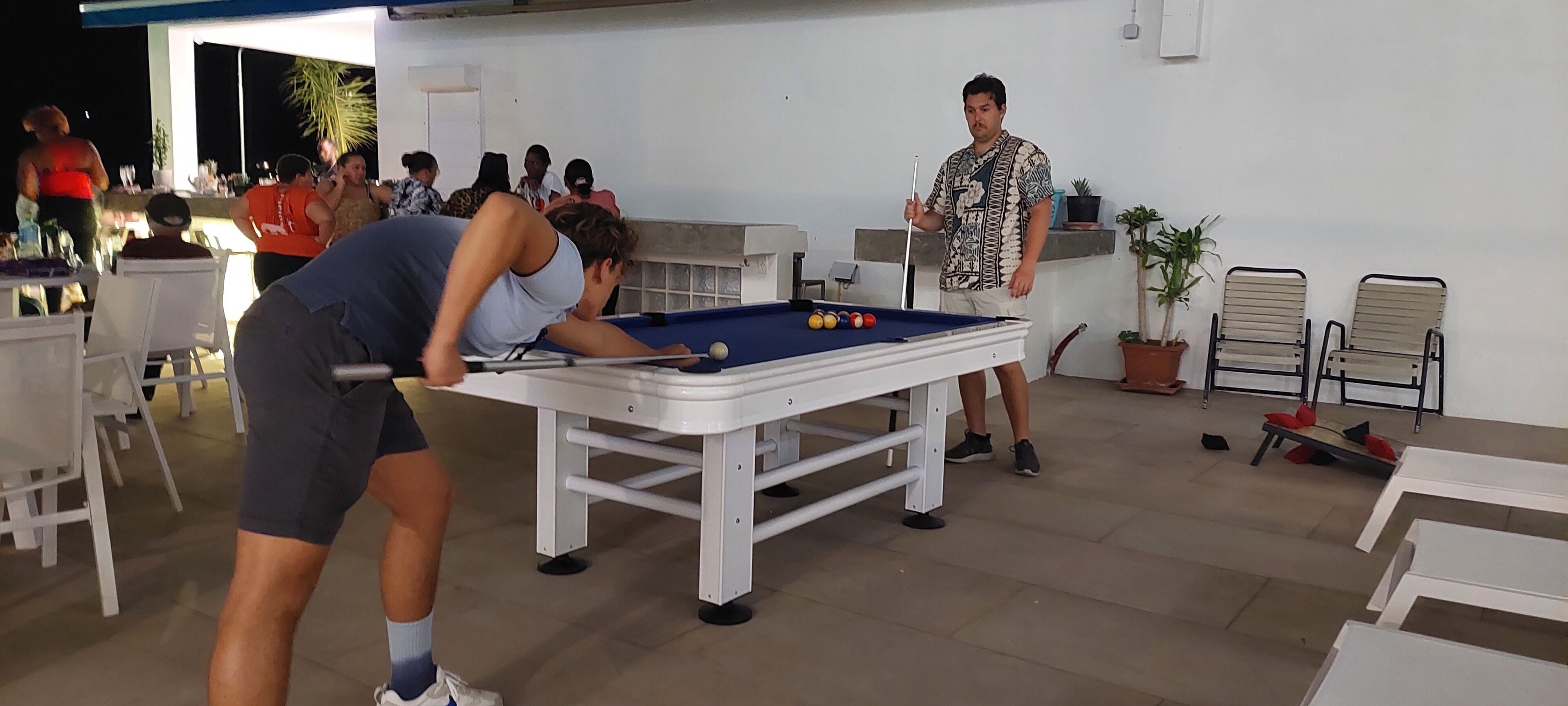 Una mesa de billar con bolas, una persona inclinada sobre ella y otra persona de pie cerca. Hay sillas y mesas alrededor, y algunas personas al fondo.