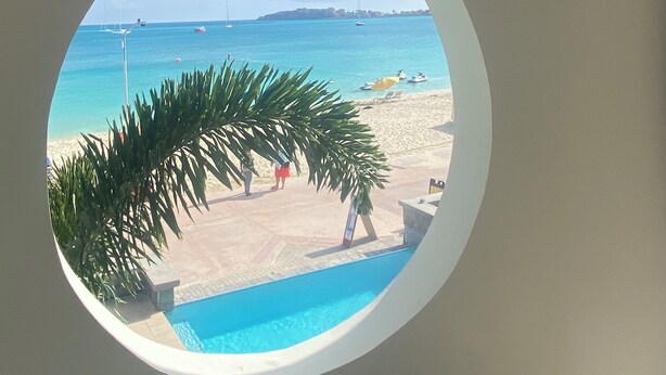 A circular window view of a beach with a swimming pool, palm trees, and a sandy area with umbrellas.