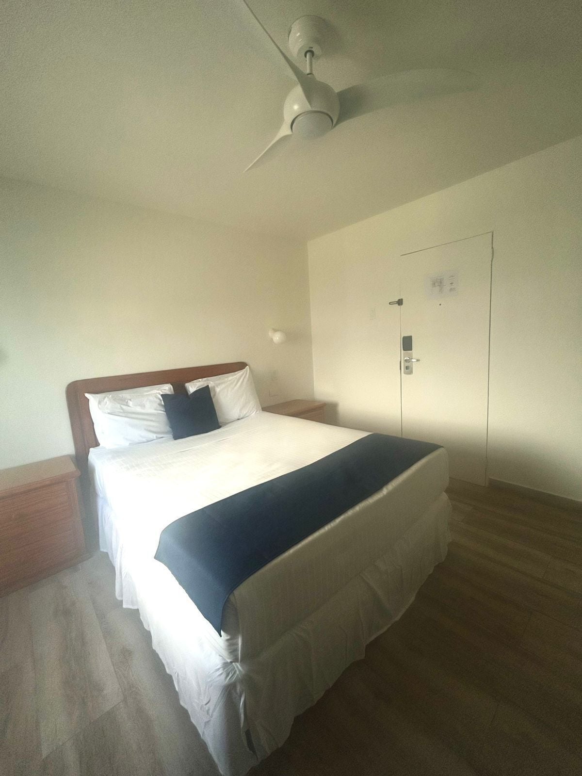 A single bed with a wooden headboard, white bedding, and a dark blue blanket. There is a ceiling fan above the bed, a wooden nightstand beside it, and a door with a lock on the adjacent wall.