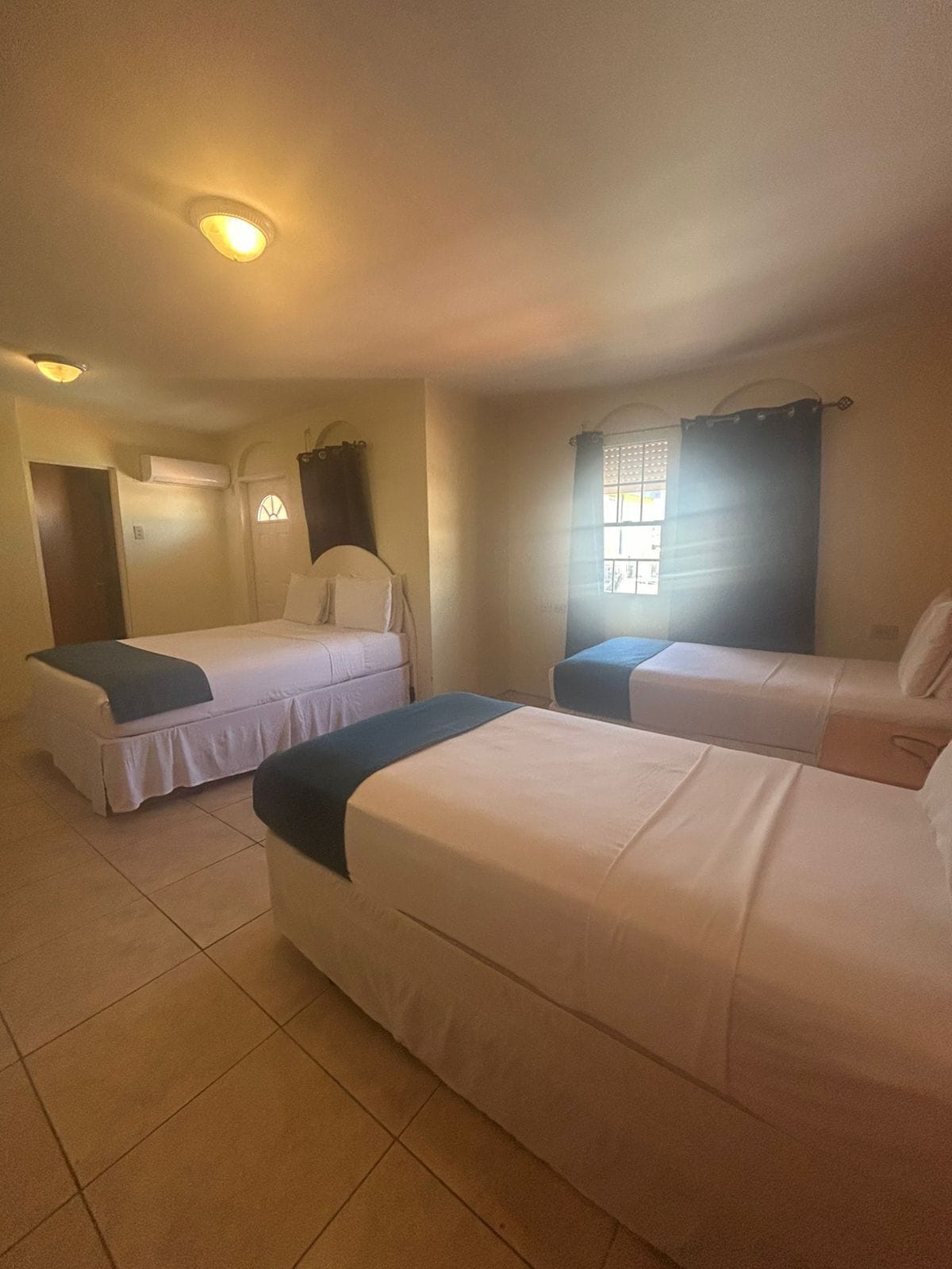 A hotel room with two single beds, a window with curtains, an air conditioning unit, and a door.