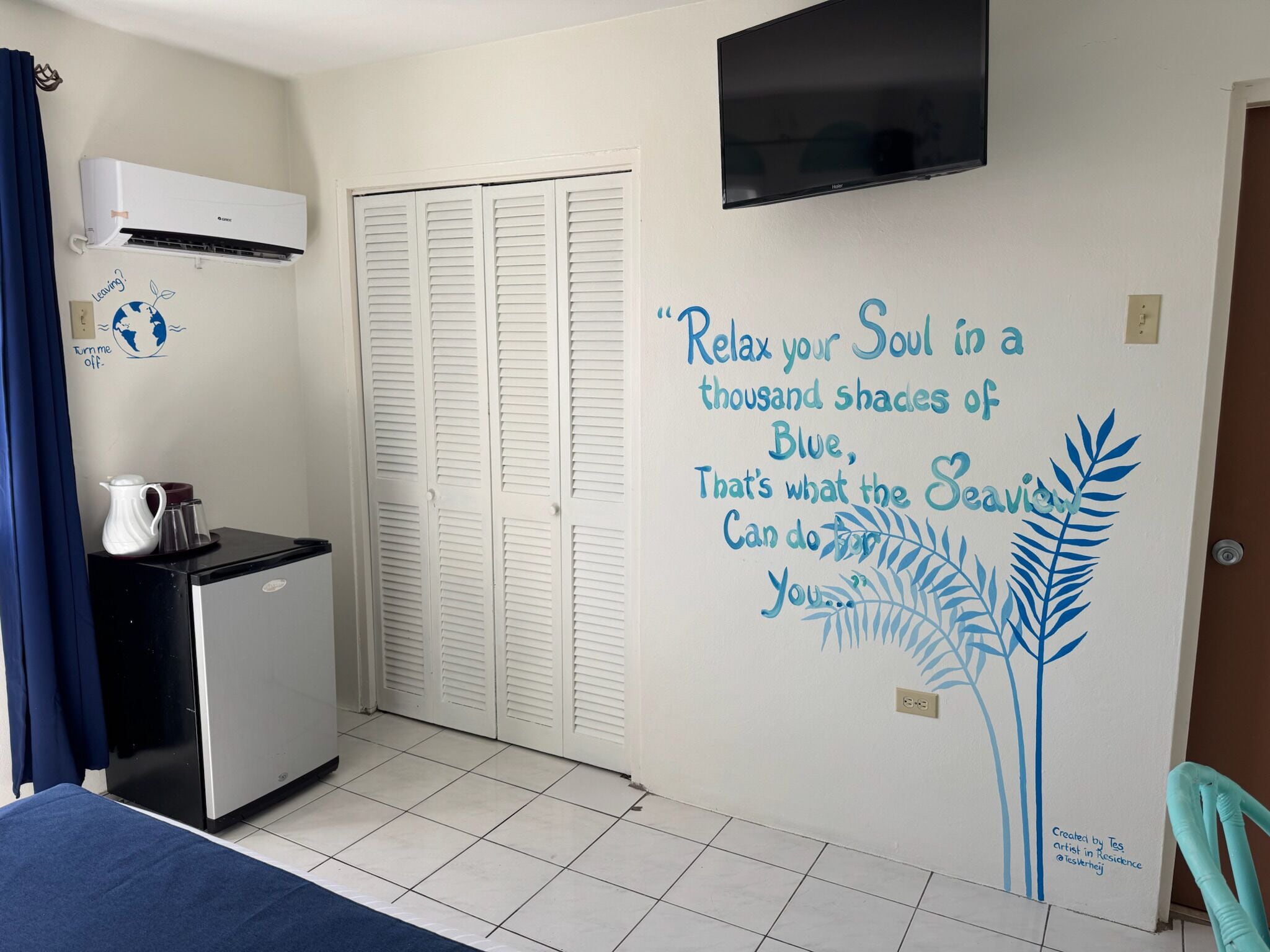 A room with a bed, a small kitchenette, a TV, and a wall with a calming quote and a leaf design.