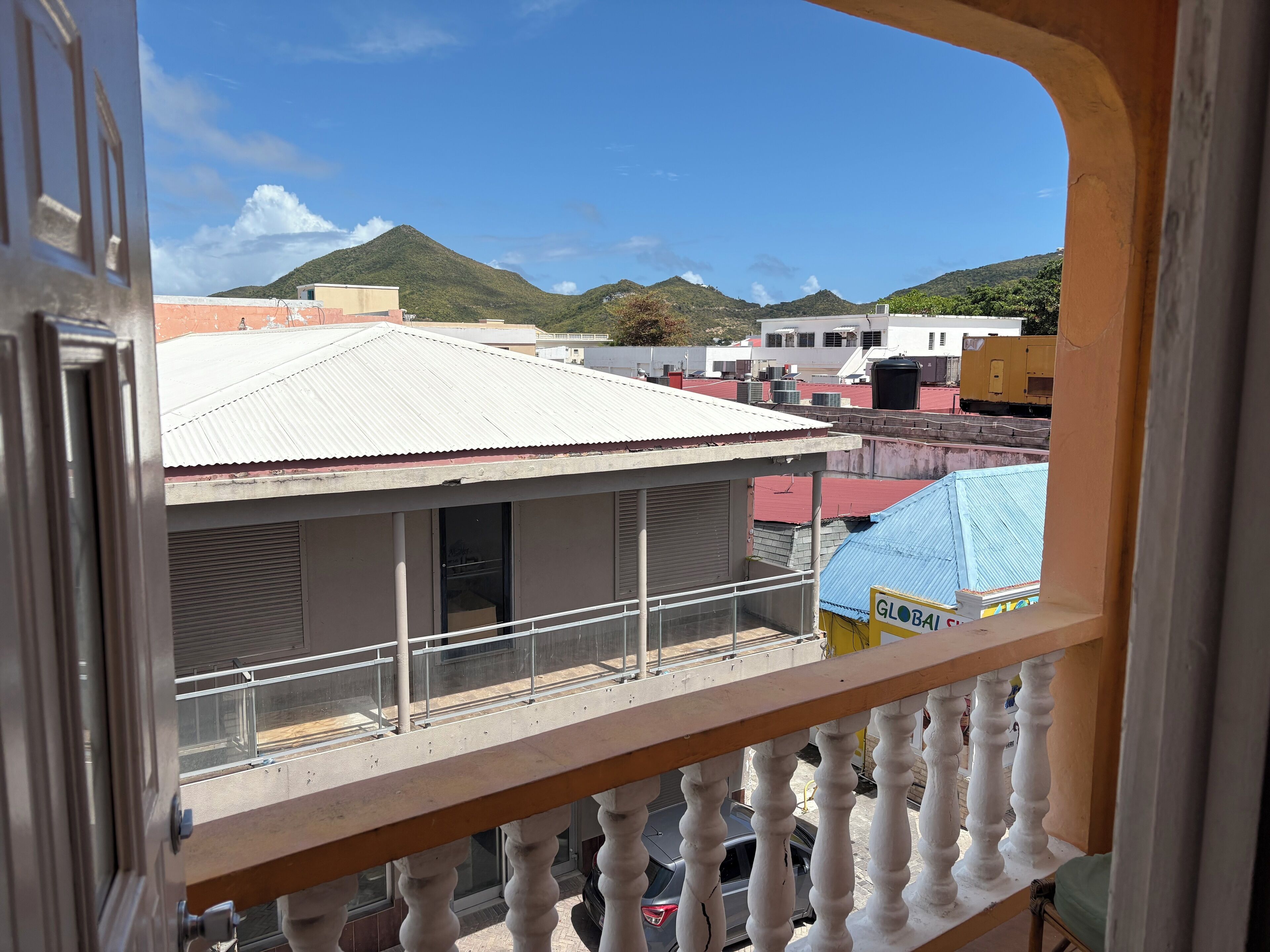 A balcony with a view of a building with a corrugated metal roof, a sign reading "GLOBAL", and a mountainous landscape in the background.