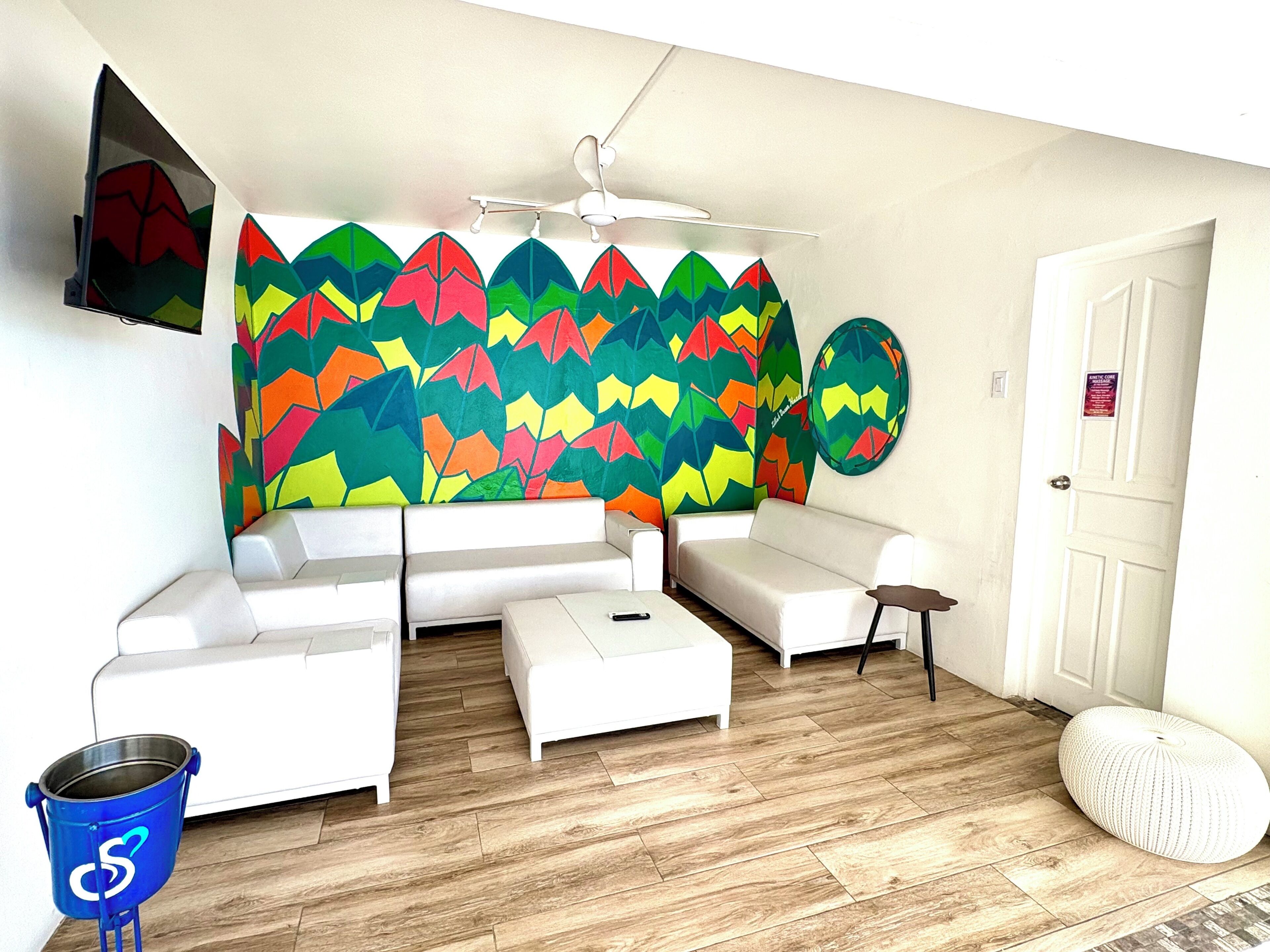A modern living room with a white sofa set, a ceiling fan, a colorful abstract wall art, and a wooden floor.