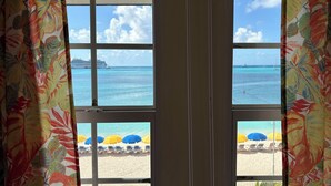 A view through a window with tropical curtains, revealing a beach with umbrellas and lounge chairs.