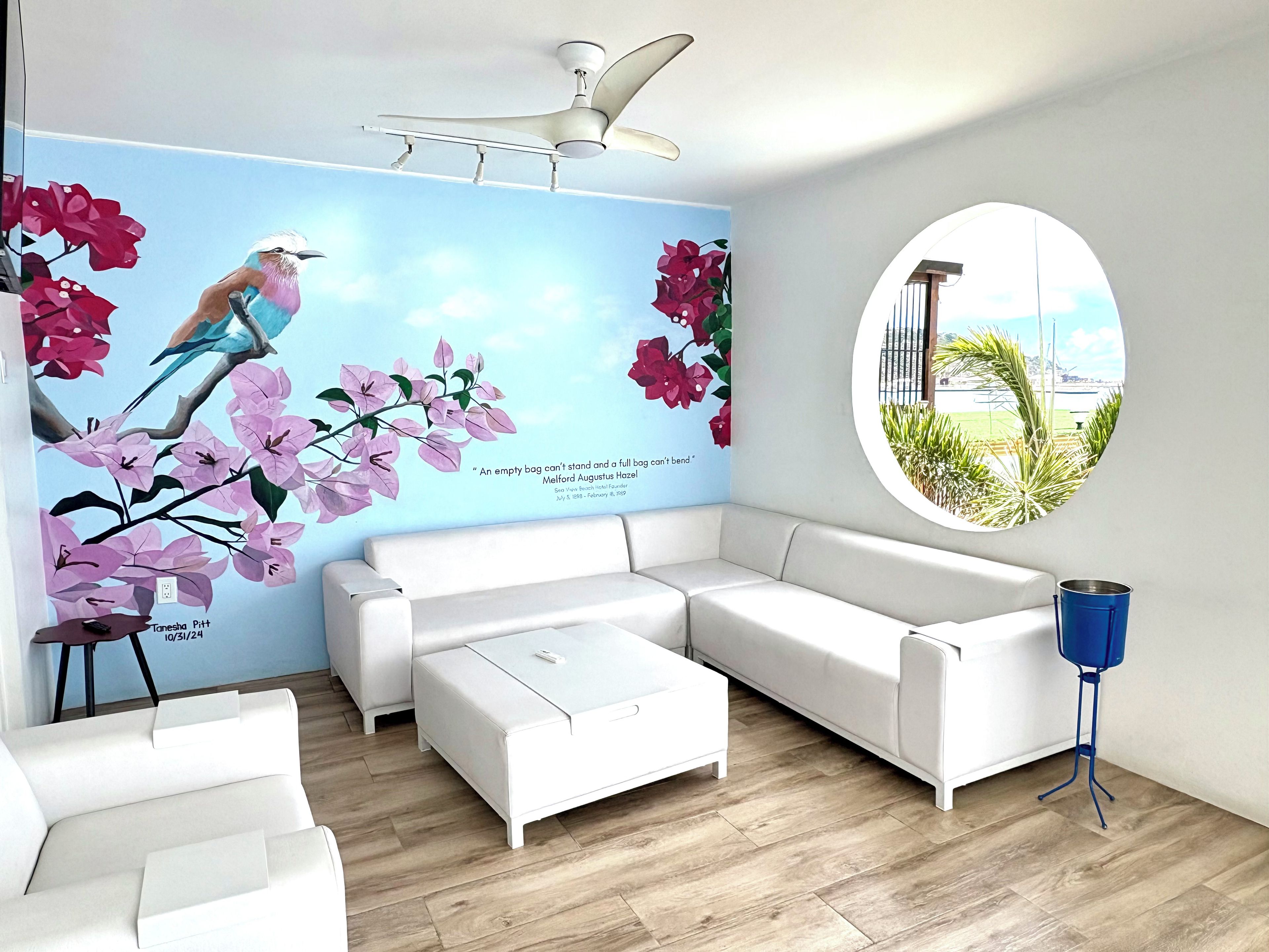 Una sala de estar moderna con un sofá seccional blanco, un ventanal redondo con vistas, un ventilador de techo y un mural de aves y flores en la pared.