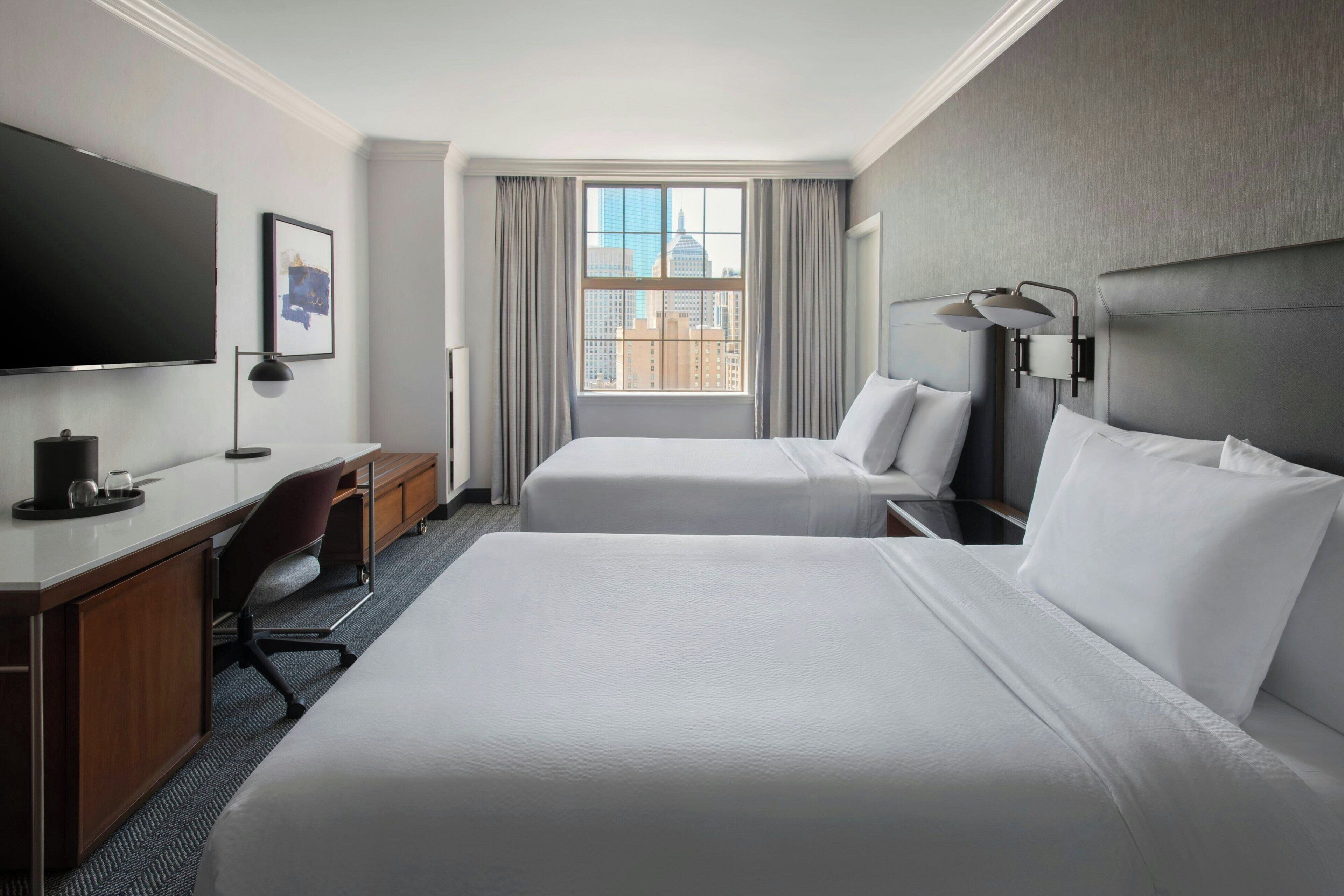Een hotelkamer met twee bedden, een bureau, een televisie en een raam met uitzicht op de stad.