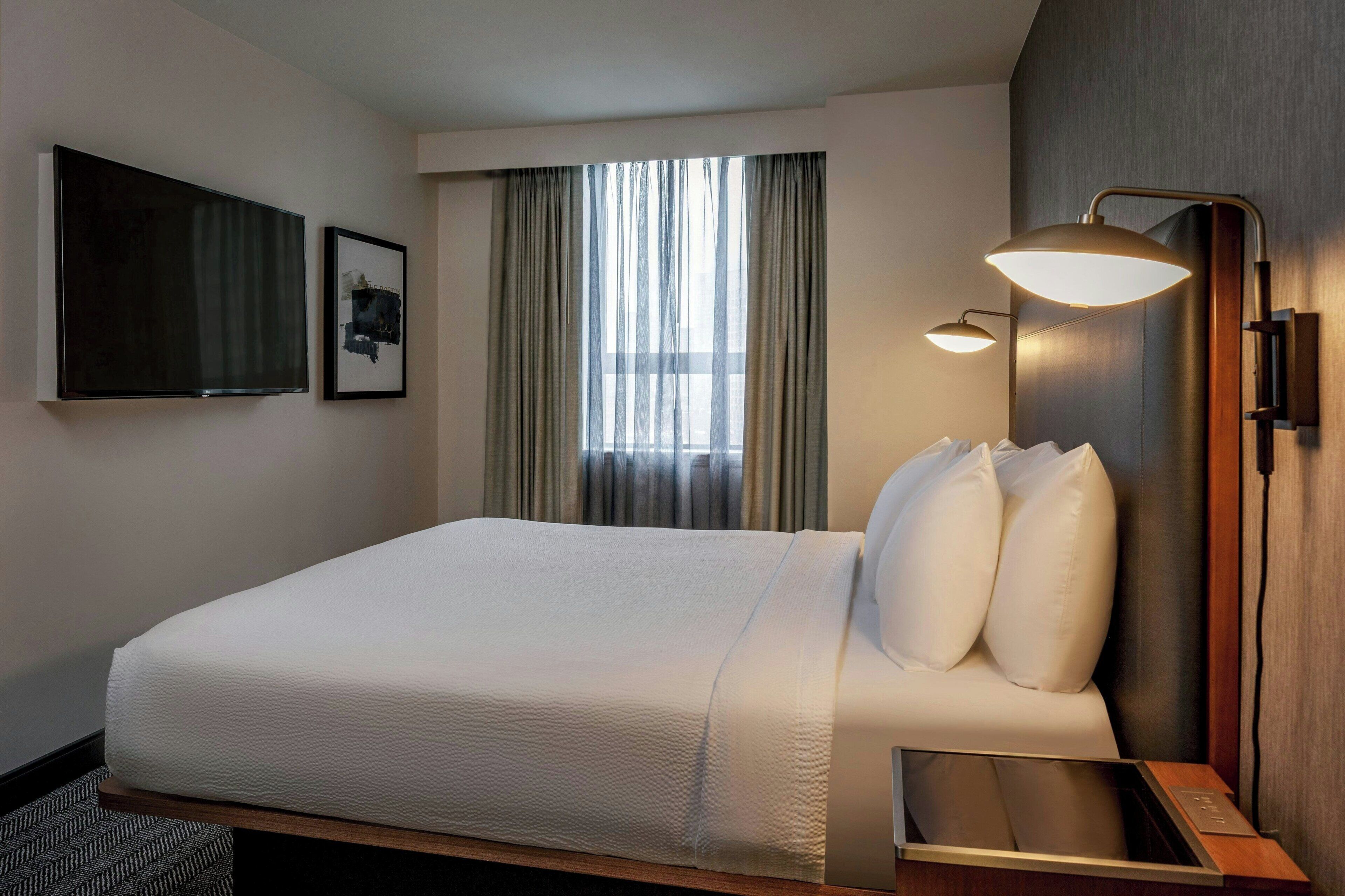 Une chambre d’hôtel avec un grand lit, une table de chevet, une lampe fixée au mur et une fenêtre avec des rideaux.