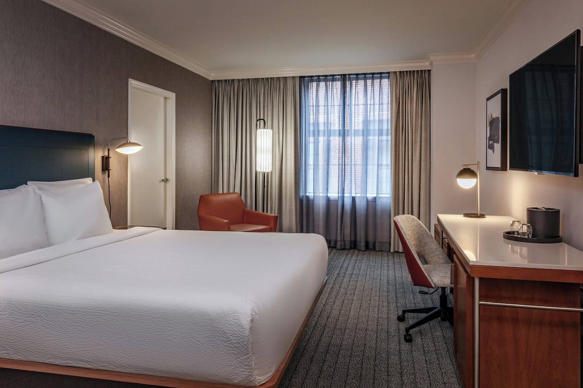 Quarto de hotel com uma cama grande, uma escrivaninha, uma cadeira, uma televisão e uma janela com cortinas.