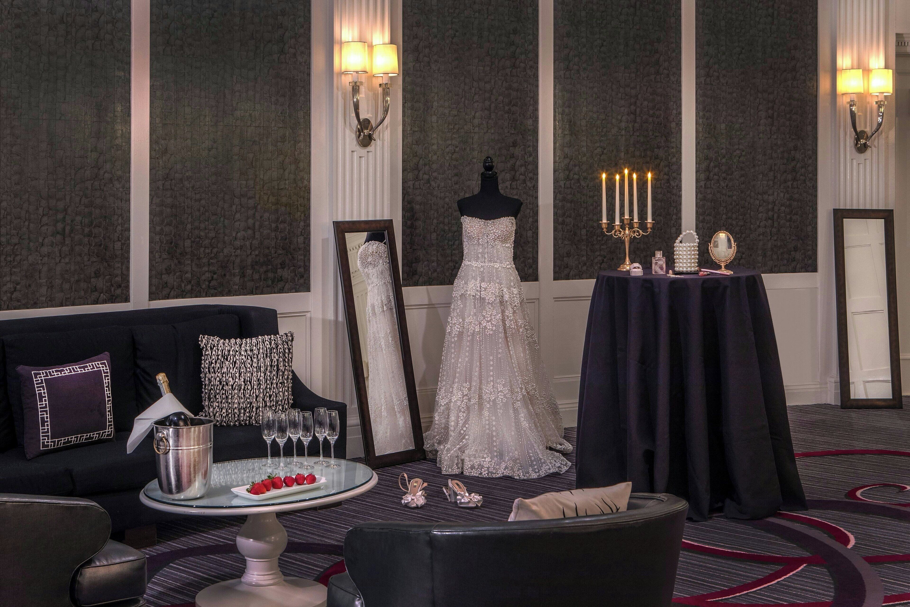 Una habitación con un sofá negro, una mesa redonda con un cubo para champán, un vestido en un maniquí y una mesa con un candelabro.