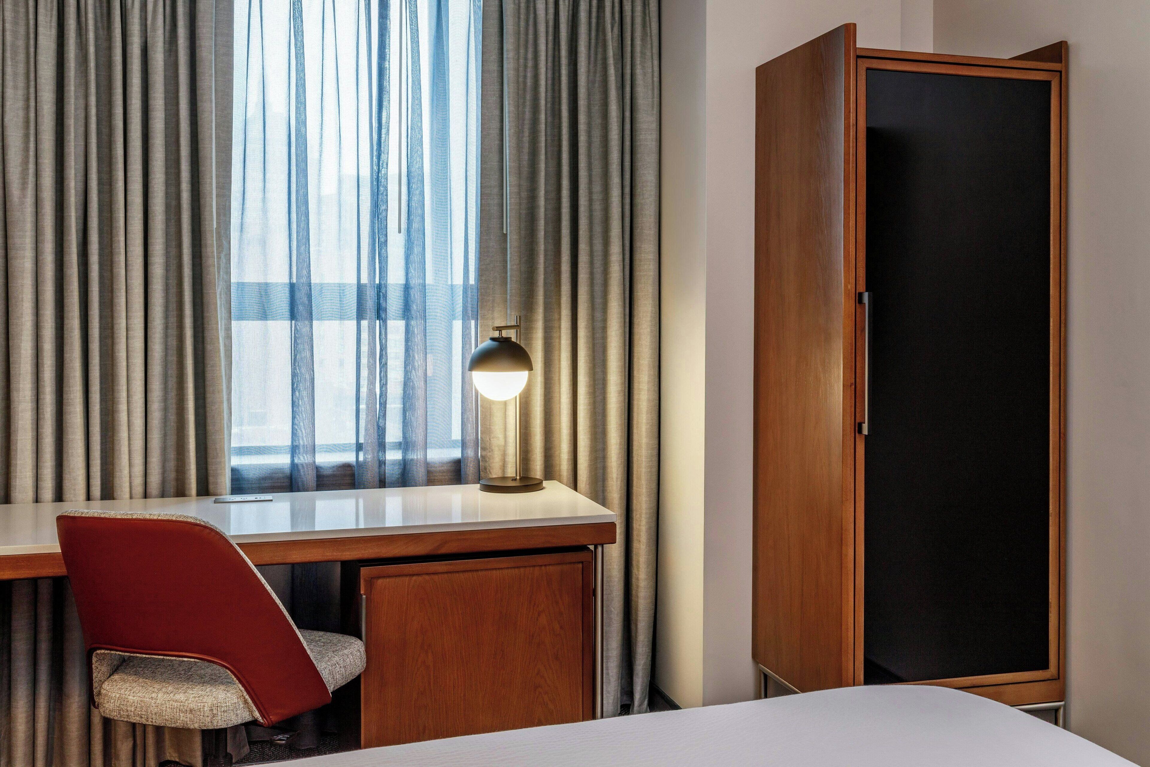 A hotel room with a desk, chair, wardrobe, and a window with curtains.